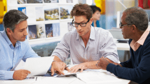 Three business professionals discussing documents and reviewing information together at a table, representing collaboration and strategic communication.